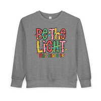 Heather-gray toddler sweatshirt with BE THE LIGHT graphic, Christmas bulbs, and Matthew 5:14. Cozy, durable fleece blend for warmth. Perfect for festive occasions.