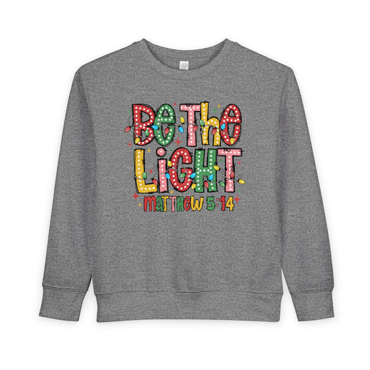 Heather-gray toddler sweatshirt with BE THE LIGHT graphic, Christmas bulbs, and Matthew 5:14. Cozy, durable fleece blend for warmth. Perfect for festive occasions.