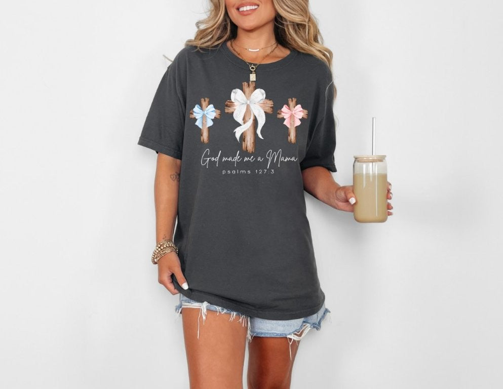 Woman wearing God Made Me a Mama oversized tee with pastel bows and wooden crosses, paired with denim shorts and accessories. Holding a drink in a jar-style cup.
