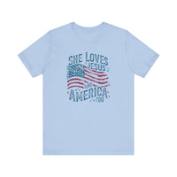 Light-blue T-shirt featuring a distressed American flag graphic with text: She Loves Jesus and America Too, epitomizing Faith Family Designs' unique and patriotic style.
