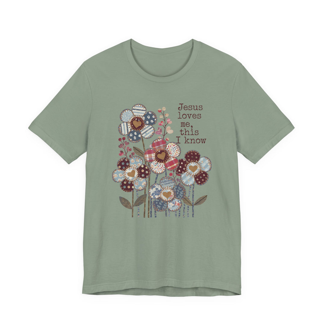 Sage-green Jesus Loves Me Tee features a patchwork floral graphic with heart centers and typewriter-style text, offering a classic fit and soft cotton comfort.