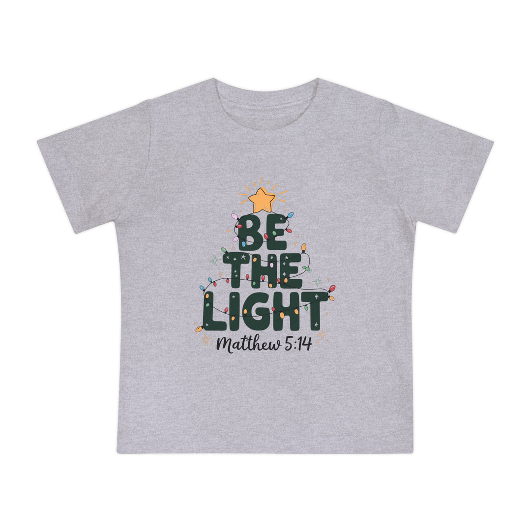 Light-gray baby tee with BE THE LIGHT in dark green, adorned with Christmas lights and sparkle accents; features Matthew 5:14 below.