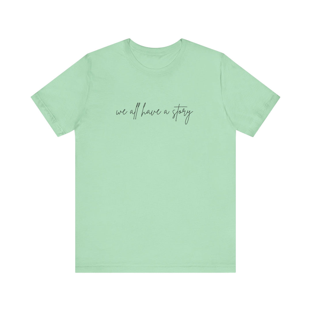 We all have a Story Tee, green with black text, soft cotton, ribbed collar, tapered shoulders, dual side seams, unisex fit, perfect for casual wear.