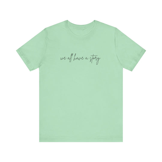 We all have a Story Tee, green with black text, soft cotton, ribbed collar, tapered shoulders, dual side seams, unisex fit, perfect for casual wear.