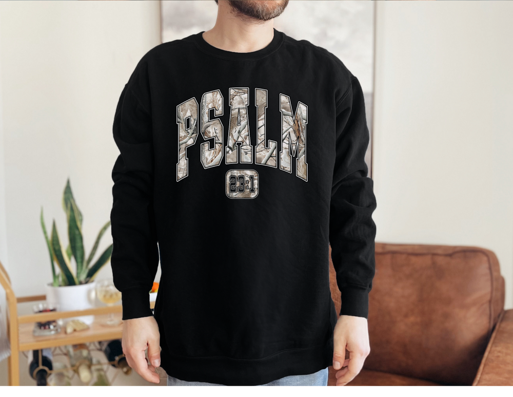Model wearing black Psalm 23 long sleeve tee in a lounge, showcasing collegiate block letters and badge. 100% ring-spun cotton, relaxed fit, ideal for casual wear.