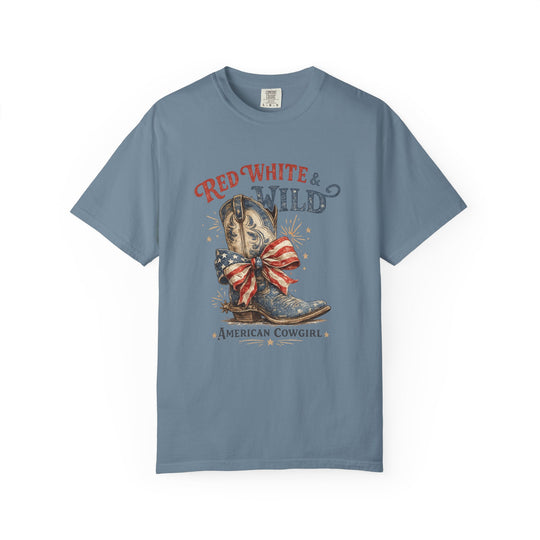 Slate-blue T-shirt with “Red White & Wild” graphic, featuring a cowboy boot with American-flag bow, labeled “American Cowgirl”. Relaxed fit, 100% ring-spun cotton.