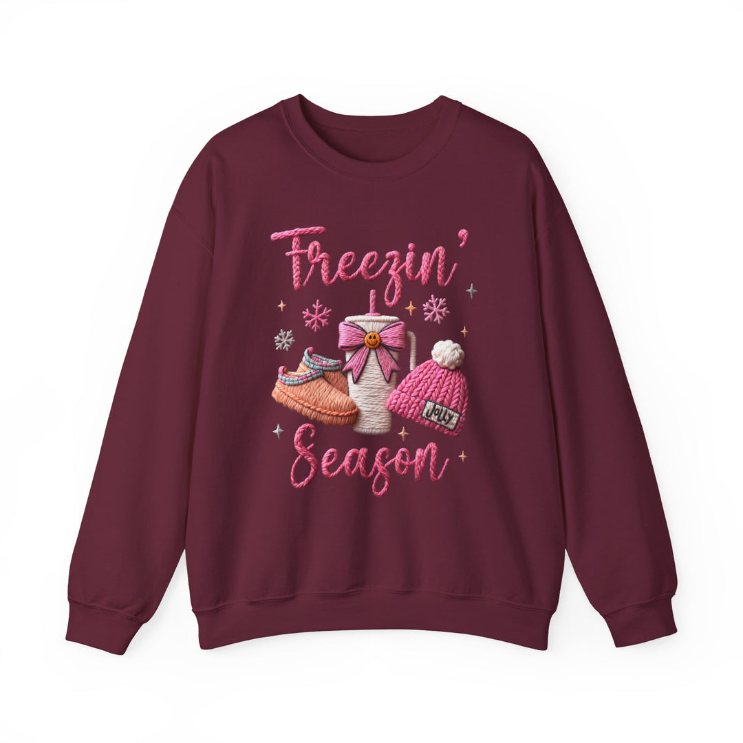 Freezin Season Crew sweatshirt features cozy motifs: Freezin’ Season in pink script, slipper shoes, to-go mug, Jolly beanie, snowflakes, and star accents.