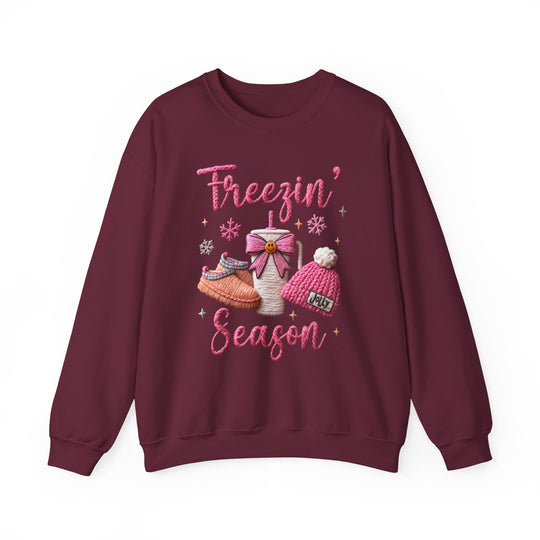 Freezin Season Crew sweatshirt features cozy motifs: Freezin’ Season in pink script, slipper shoes, to-go mug, Jolly beanie, snowflakes, and star accents.
