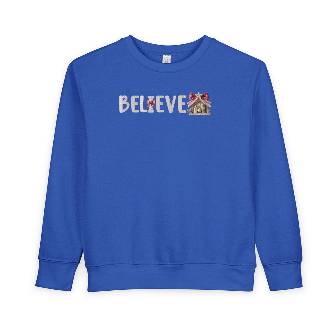 Royal-blue toddler crewneck sweatshirt with BELIEVE in white knit-look font, featuring a nativity scene as the final E and a red bow on the I.