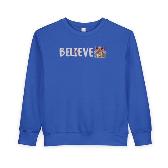 Royal-blue toddler crewneck sweatshirt with BELIEVE in white knit-look font, featuring a nativity scene as the final E and a red bow on the I.