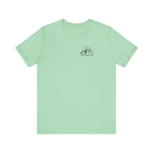 A green tee with a mountain logo, embodying the Faith Can Move Mountains Tee by Worlds Worst Tees. Unisex, 100% cotton, retail fit, tear away label, and ribbed knit collars for a beloved favorite.