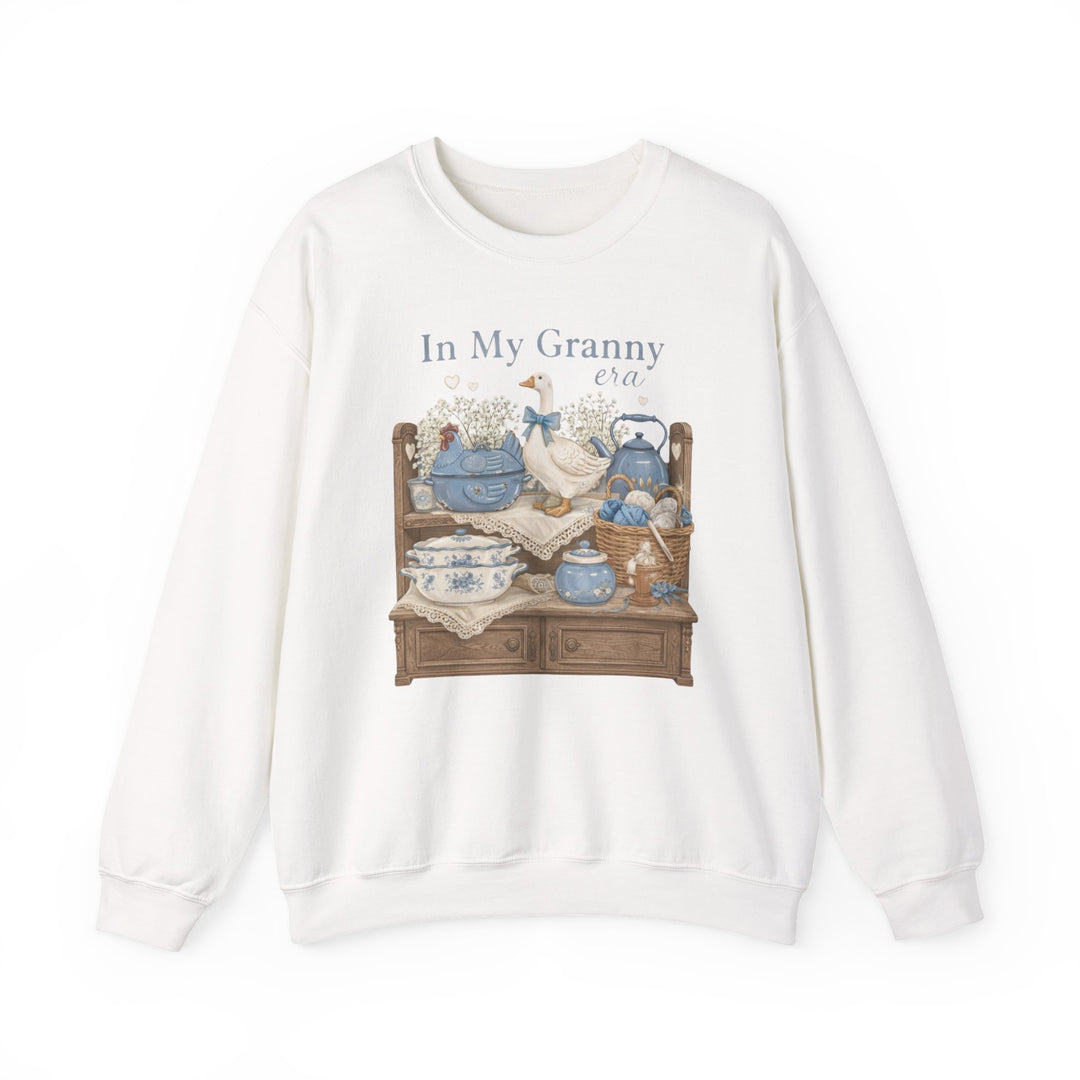 In My Granny Era Crew: White sweatshirt with whimsical hutch graphic, blue china, a goose figurine, and knitting basket, offering cozy comfort with a polyester-cotton blend.