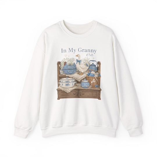 In My Granny Era Crew: White sweatshirt with whimsical hutch graphic, blue china, a goose figurine, and knitting basket, offering cozy comfort with a polyester-cotton blend.