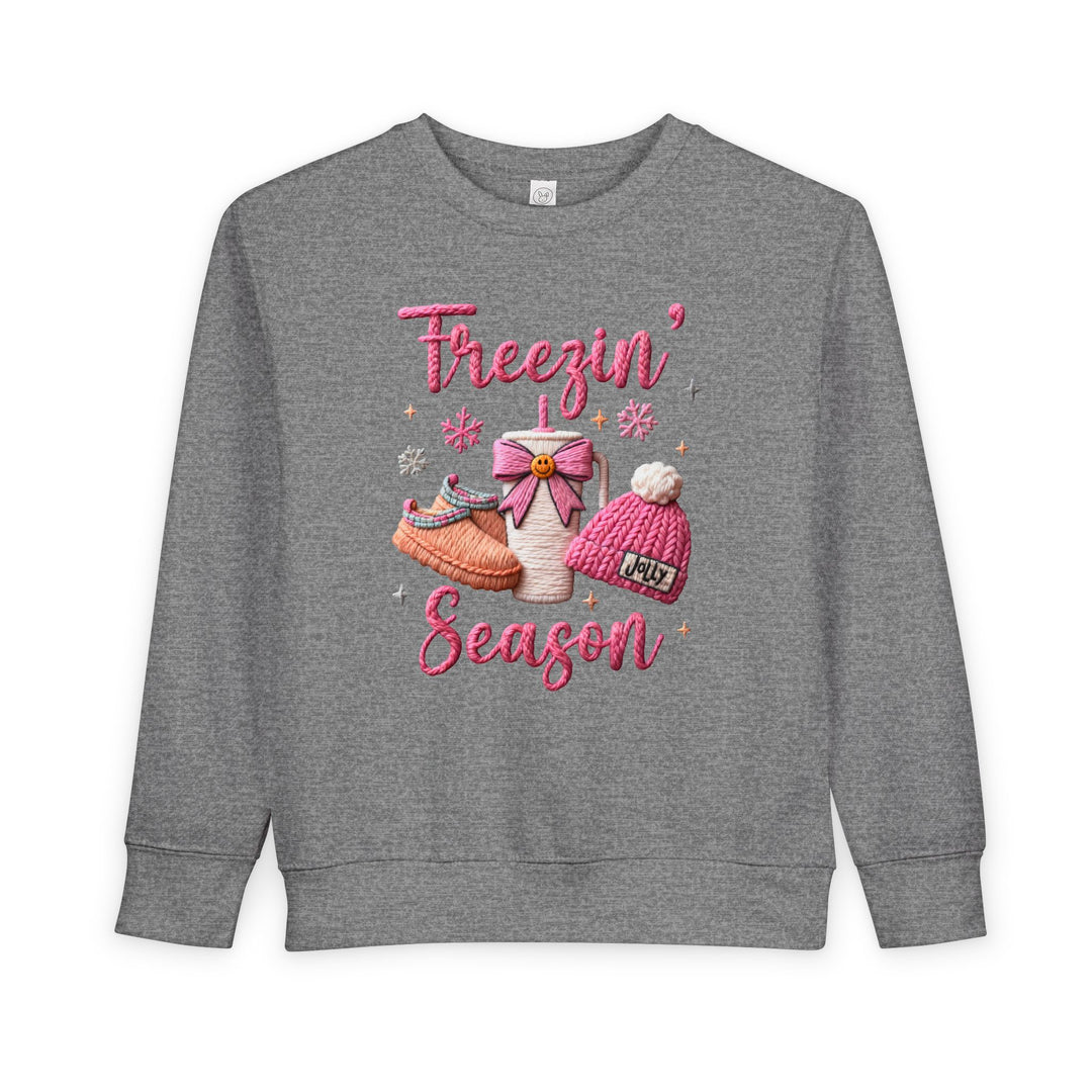 Freezin Season Toddler Crew: Heather-gray sweatshirt with playful winter motifs—pink lettering, hot-drink cup, slippers, beanie, and snowflakes—crafted for warmth and comfort.