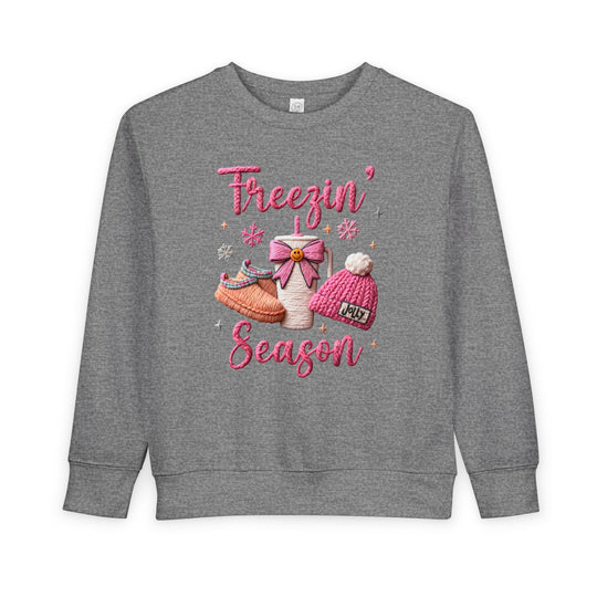Freezin Season Toddler Crew: Heather-gray sweatshirt with playful winter motifs—pink lettering, hot-drink cup, slippers, beanie, and snowflakes—crafted for warmth and comfort.
