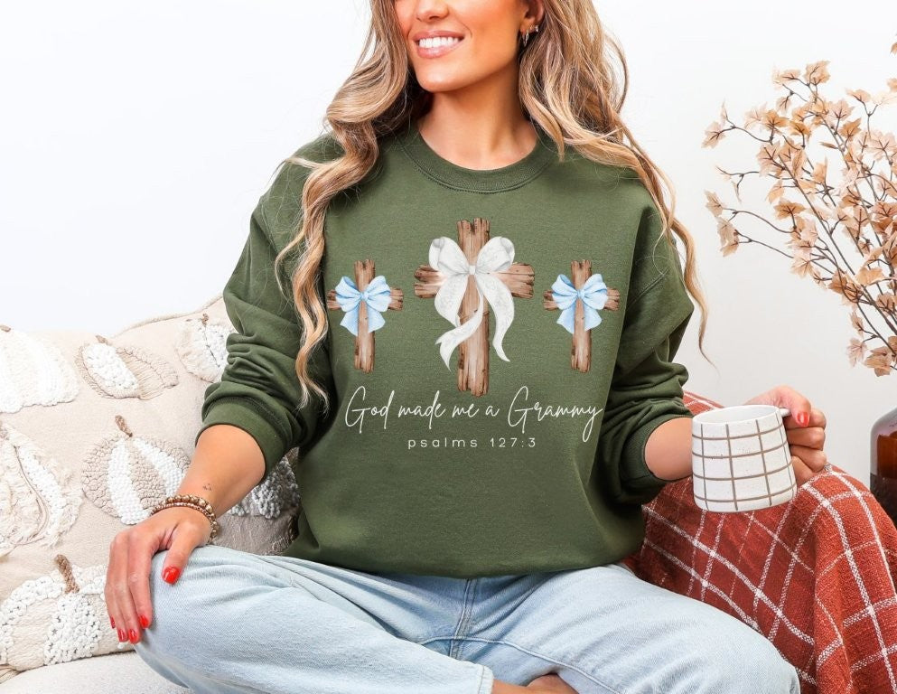 Woman wearing God Made Me a Grammy forest-green sweatshirt with cross design, holding a patterned mug on a couch.