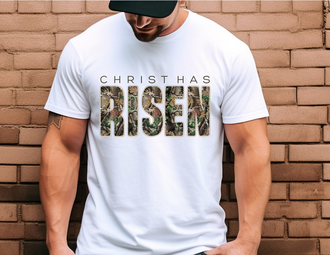 Man wearing Christ has Risen tee with camouflage RISEN letters, standing against a brick wall.