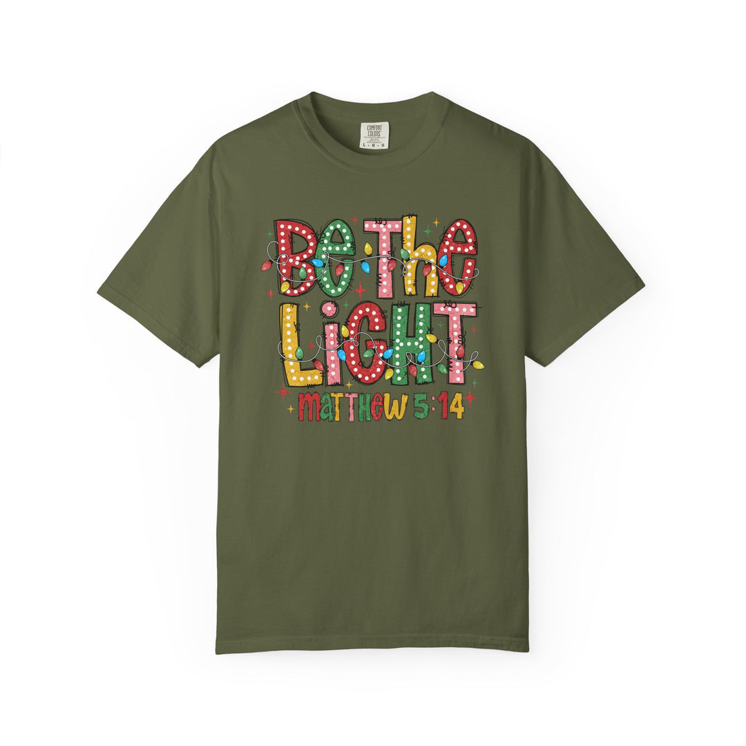 Be the Light Tee featuring Be the Light in multicolored letters and Matthew 5:14, with cartoon string-lights on an olive-green Comfort Colors T-shirt.