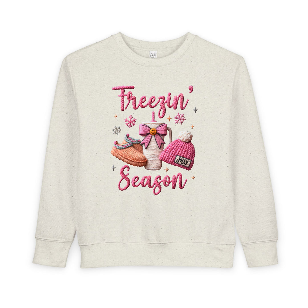 Toddler sweatshirt with Freezin’ Season in pink script and cozy motifs: slippers, thermos with bow, and pom-pom beanie. Designed for warmth and comfort.
