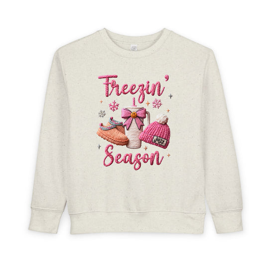 Toddler sweatshirt with Freezin’ Season in pink script and cozy motifs: slippers, thermos with bow, and pom-pom beanie. Designed for warmth and comfort.