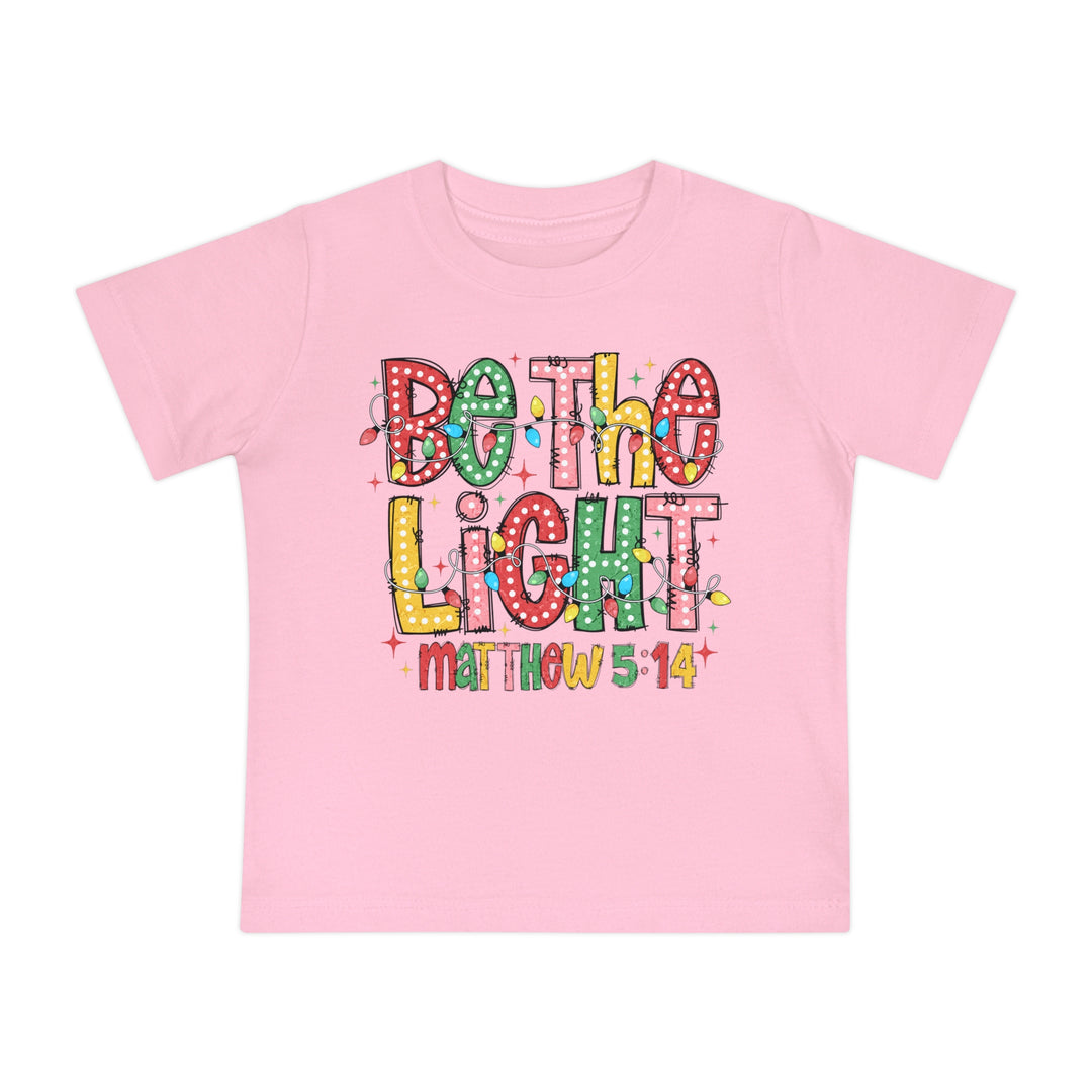 Pink baby tee with Be the Light and Matthew 5:14 in festive lettering, featuring string lights, from Faith Family Designs.