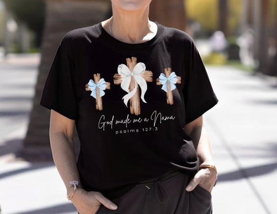 Person wearing a God Made Me a Nana tee, featuring three wooden crosses with bows, hands in pockets, outdoors.