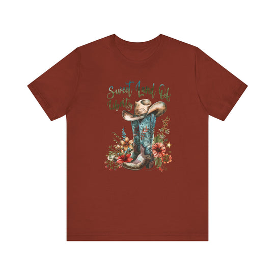 A red shirt featuring a cowboy hat and flowers, embodying a classic Americana vibe. Unisex jersey tee with ribbed knit collars, taping on shoulders, and 100% Airlume combed cotton for comfort. Sweet Land of Liberty Tee by Worlds Worst Tees.