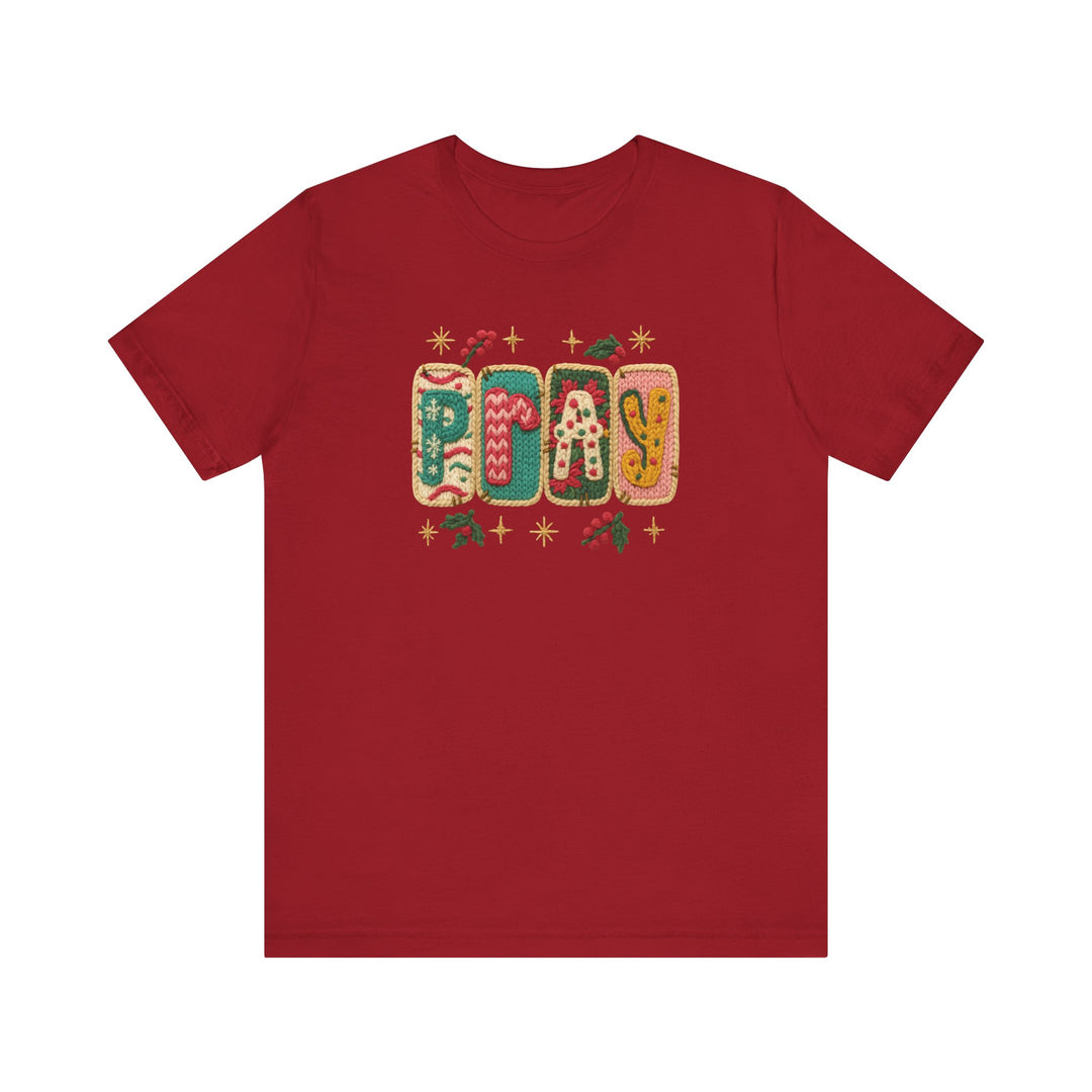 Unisex Pray Tee with a holiday-themed PLAY graphic, featuring knitted pillow-style letters and festive motifs, on a soft cotton red T-shirt.