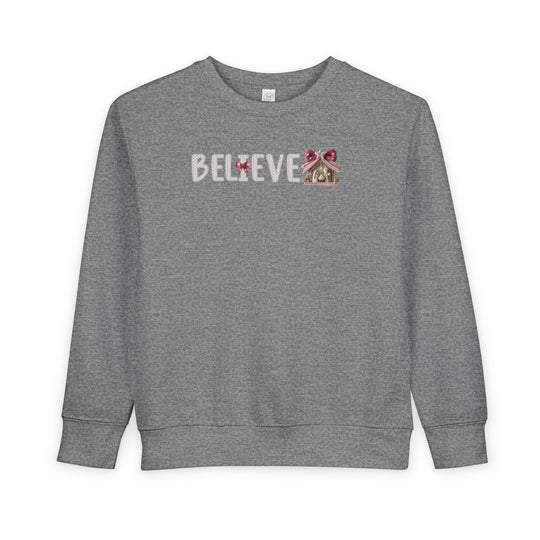Heather-gray toddler crewneck with BELIEVE in white stitch; gingham bows adorn letters, ending in a nativity scene under a shelter with a star.