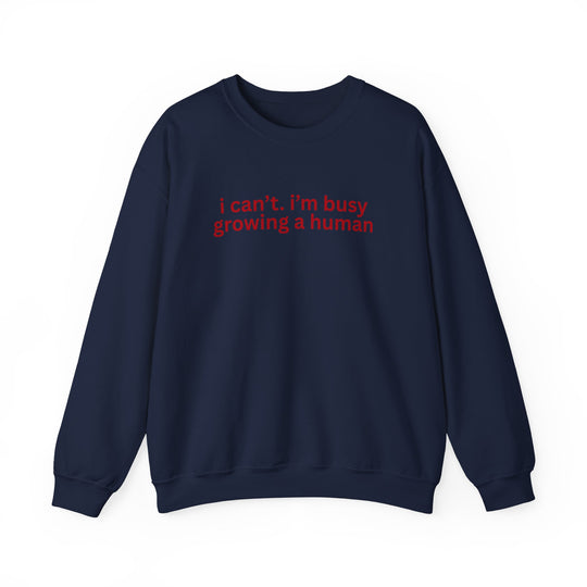 Navy-blue crewneck sweatshirt with red text, i can’t. i’m busy growing a human. Laid flat, showcasing its cozy design and durable stitching.