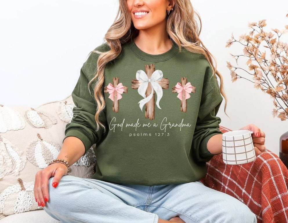 Woman in God Made Me a Grandma sweatshirt with wooden cross design, holding a mug, seated comfortably, showcasing the sweatshirt's casual, cozy fit.
