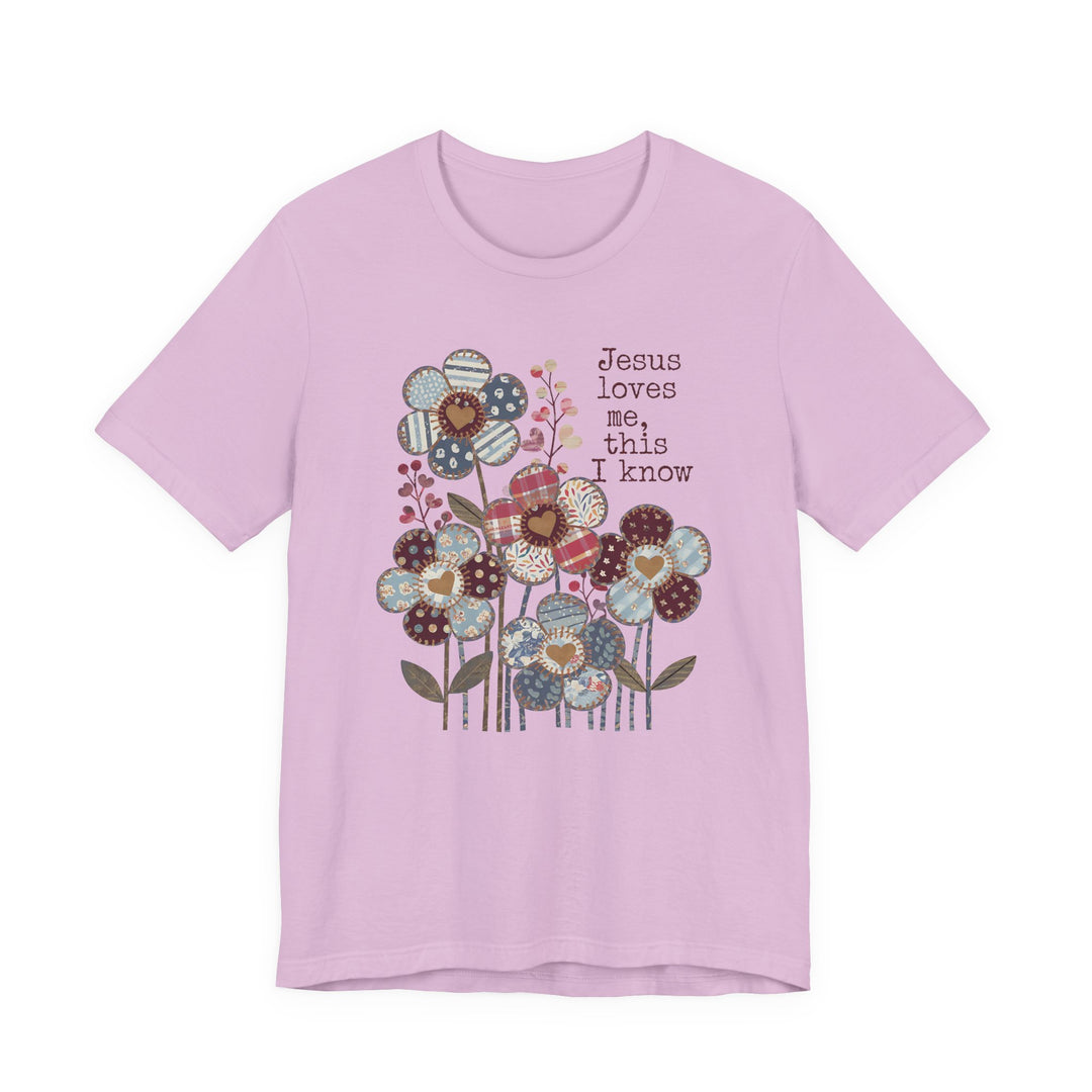 Light pink T-shirt featuring whimsical patchwork flowers and Jesus loves me, this I know text, offering a soft, comfortable fit from Faith Family Designs.