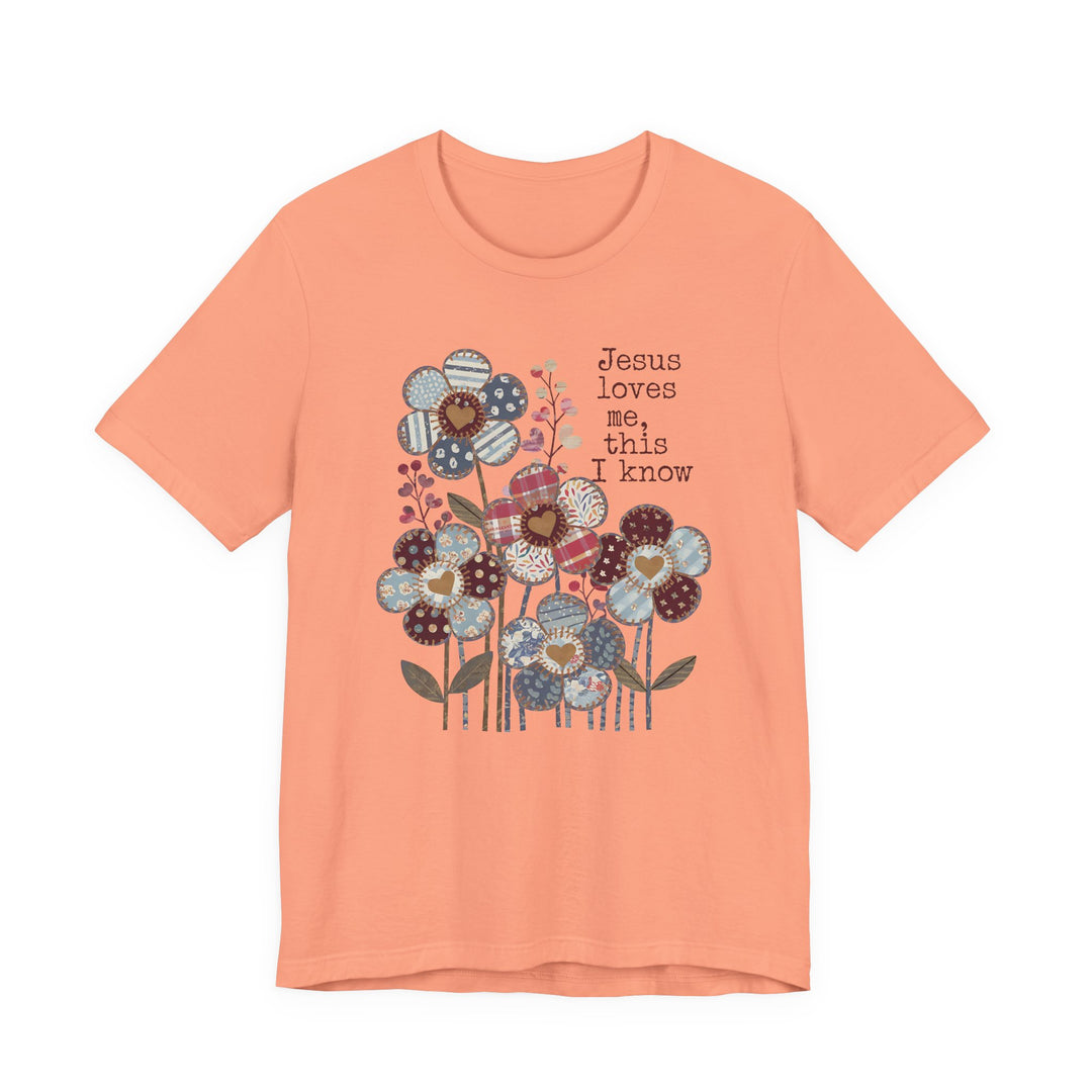 Peach-colored Jesus Loves Me Tee with patchwork flowers and typewriter-style text, embodying a soft, classic fit from Faith Family Designs.