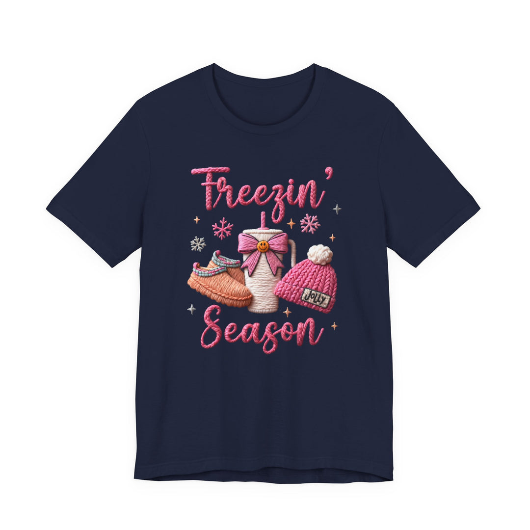 Navy-blue T-shirt with Freezin' Season in pink yarn lettering, featuring a winter hat, slippers, and steaming cup graphic, adorned with snowflakes for a festive touch.