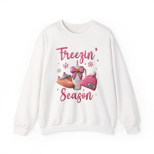 Freezin Season Crew: White crew-neck sweatshirt featuring winter-themed graphics with a steaming mug, slippers, beanie, and snowflakes, perfect for cozy comfort.