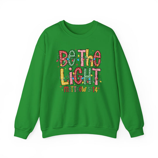 Green crewneck sweatshirt featuring a festive Be the Light graphic with colorful lights and Matthew 5:14, reflecting a playful holiday style.