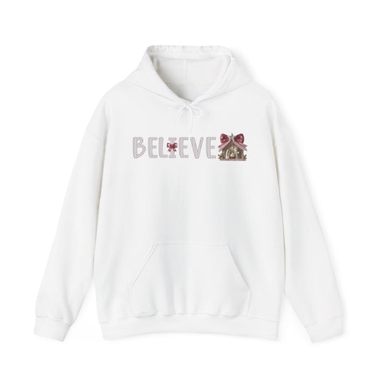White Believe hoodie with a knit-style font, red bow detail, and nativity-stable graphic. Features a kangaroo pocket and matching drawstring hood for warmth and style.