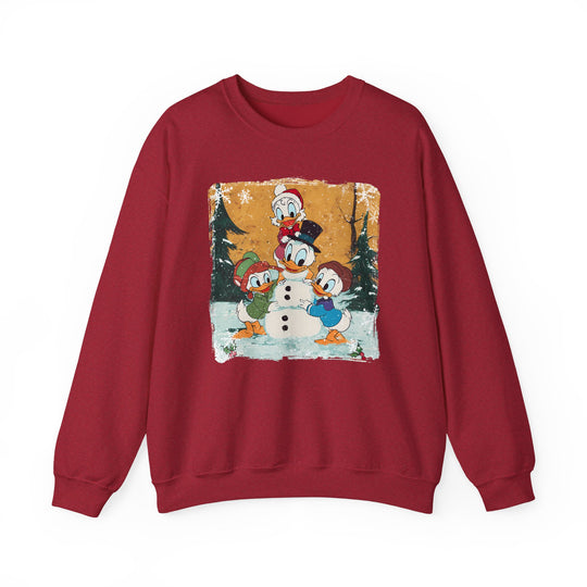 Red crew-neck sweatshirt featuring three ducks in winter attire building a snowman amidst a snowy pine-tree backdrop, offering cozy comfort for holiday festivities.