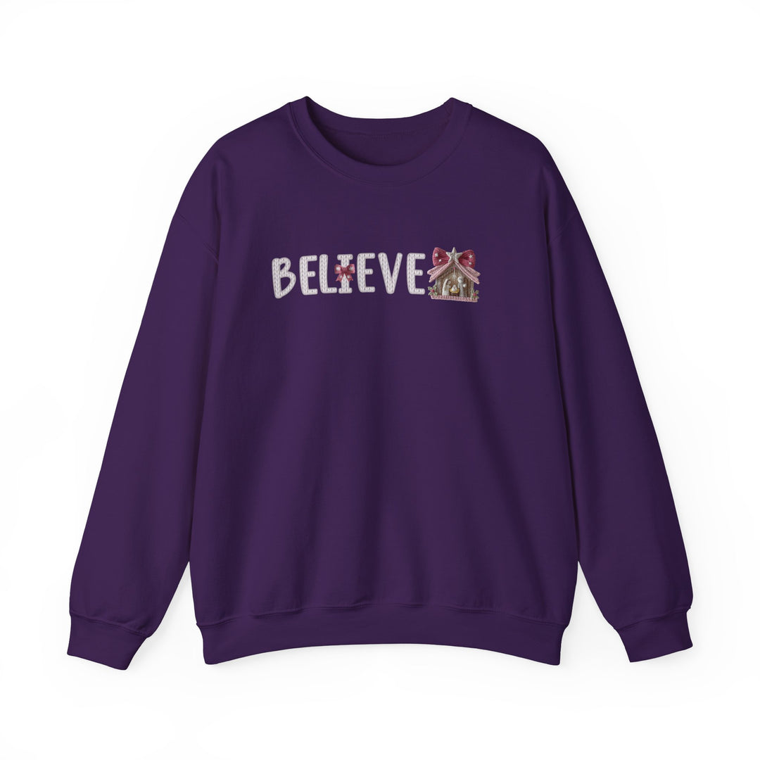 Purple crew-neck sweatshirt with BELIEVE in knit-style font; red bow replaces I, gingerbread-house ornament replaces final E, offering cozy, festive appeal.