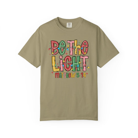 Light-tan 'Be the Light' T-shirt featuring festive bulb letters and Christmas lights graphic, referencing Matthew 5:14, from Faith Family Designs.