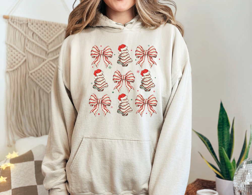 Person wearing the Cookies and Bows Hoodie, featuring festive graphics of bows and Christmas trees with Santa hats, set against a cozy home backdrop.