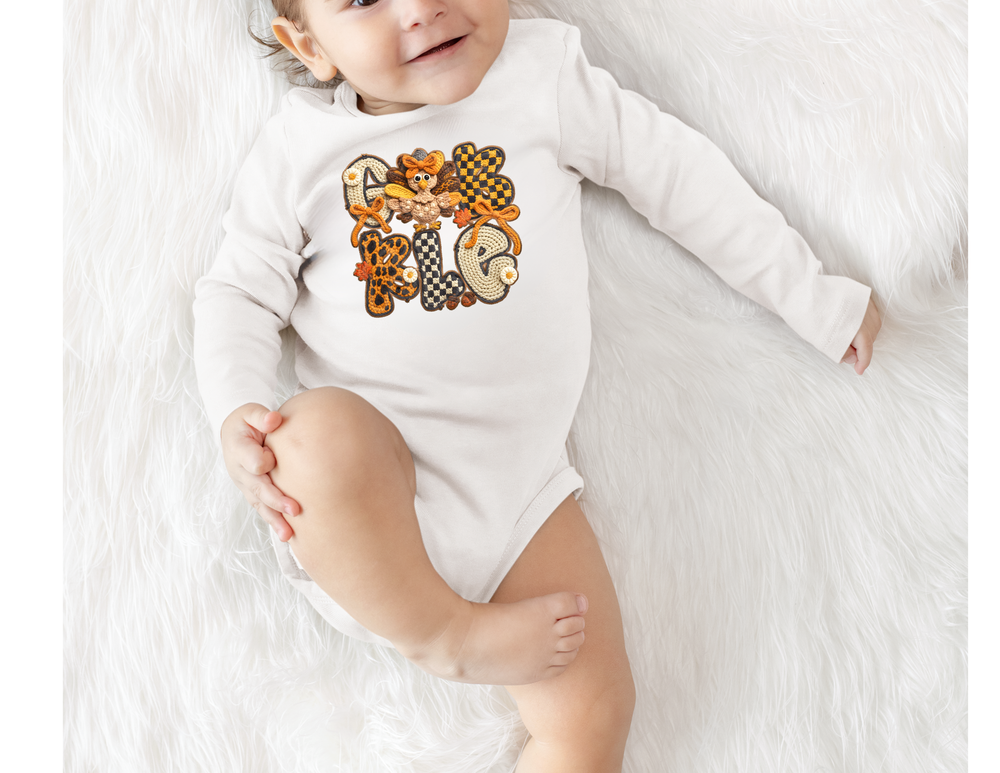 Baby in Gobble Onesie featuring autumn-themed embroidery with a cartoon turkey, lying on a fluffy surface. Snap closures for easy changing.