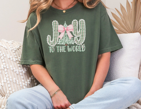 Person on sofa wearing Joy to The World Tee with festive cactus and Christmas tree design, paired with jeans, showcasing a casual holiday vibe.