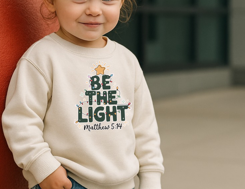 Toddler wearing cream Be The Light sweatshirt with Christmas tree graphic, designed by Faith Family Designs for warmth and style.