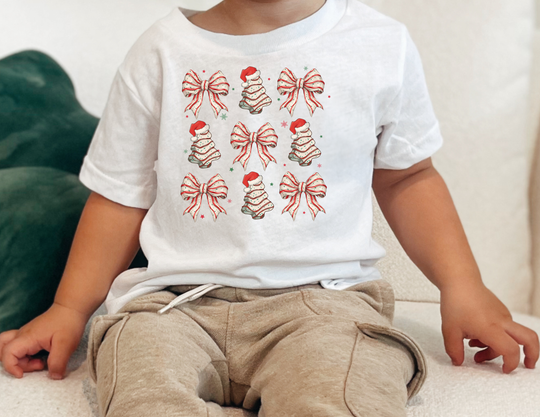 Child wearing Cookies and Bows Toddler Tee with Christmas-themed pattern, seated. White tee features gift bows and Santa-hat trees; paired with tan drawstring pants.
