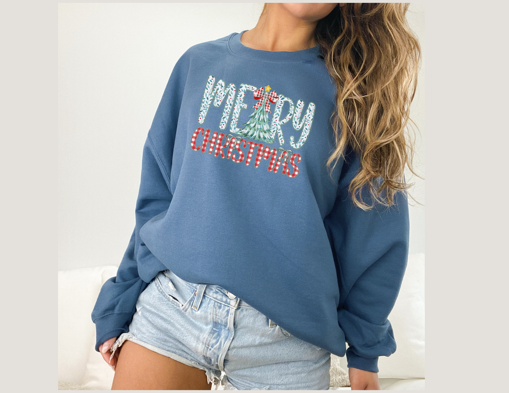 Person wearing a slate-blue Tree Christmas Crew sweatshirt with festive lettering and a Christmas tree design, paired with light-wash, frayed denim shorts.