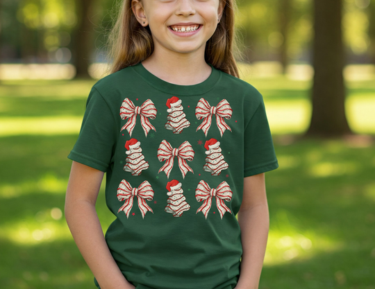 Young girl smiles in park wearing Cookies and Bows Kids Tee, featuring festive motifs of bows and bells with Santa hats on a dark green background.