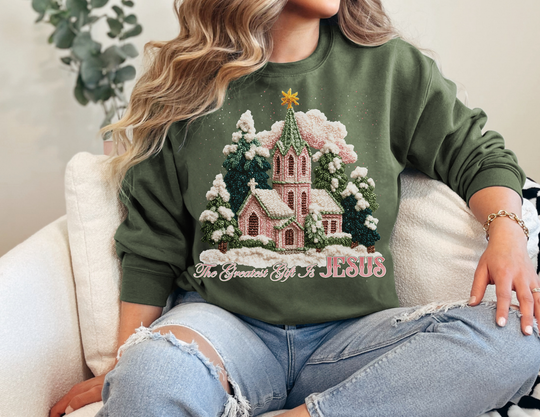 Person in a forest-green Gift is Jesus Crew sweatshirt with a holiday scene, seated on a light sofa, wearing ripped light-wash jeans.
