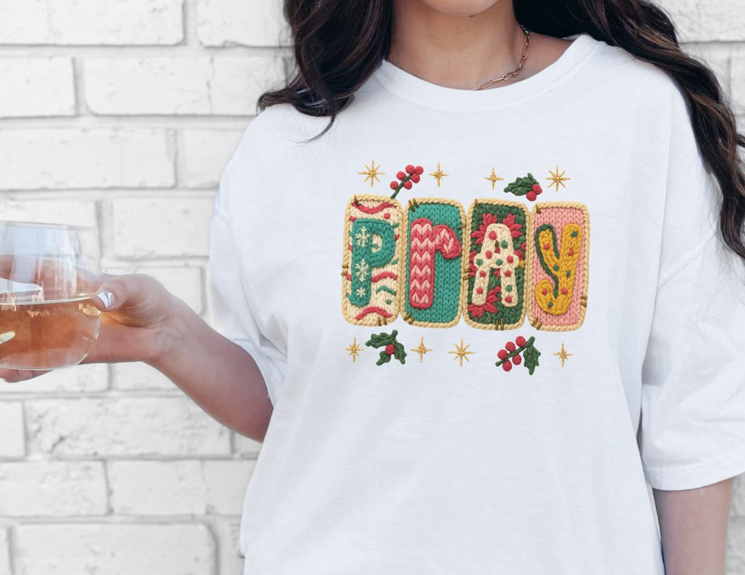 Person wearing Pray Tee with holiday-style knit print, holding a stemless wine glass, standing against a white brick wall.