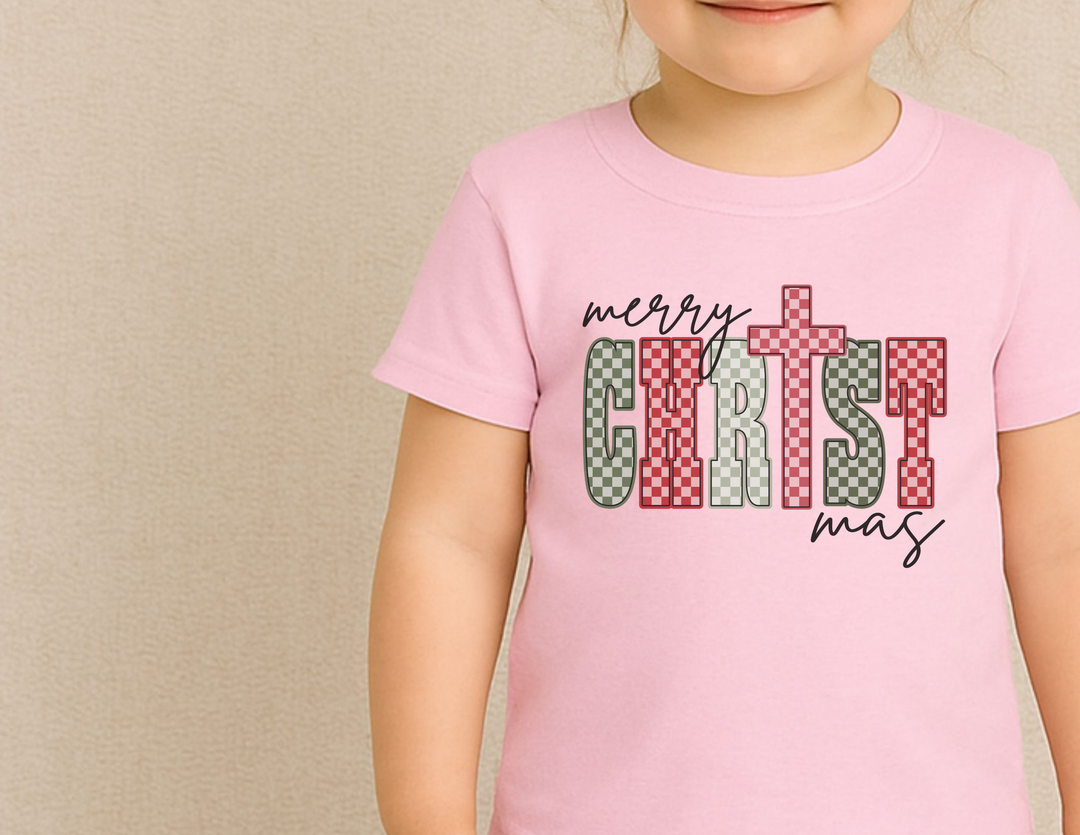 Child wearing a light-pink merry CHRIST mas Christmas Baby Tee with checkerboard lettering, featuring a cross-shaped T. Ideal for festive, comfortable wear.